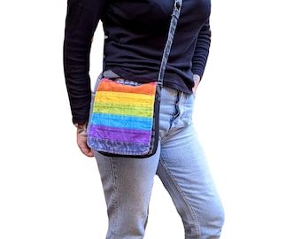 May include: A rainbow striped crossbody bag with a denim strap. The bag is worn by a person wearing a black long-sleeved shirt and blue jeans.