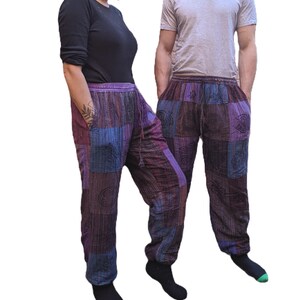 May include: Two people wearing purple, blue and brown patchwork trousers with embroidered symbols. The trousers have an elastic waistband and a drawstring.