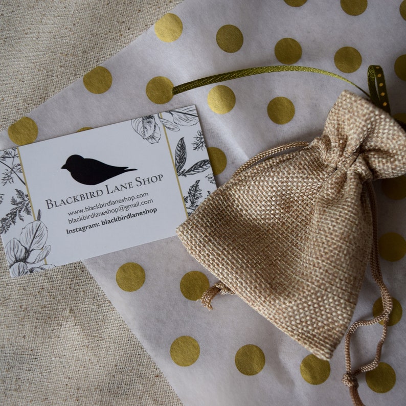 May include: A small burlap bag with a drawstring closure, sitting on white tissue paper with gold polka dots. A business card with the text "Blackbird Lane Shop" is also visible. The card includes the website address "www.blackbirdlaneshop.com", email address "blackbirdlaneshop@gmail.com", and Instagram handle "blackbirdlaneshop".