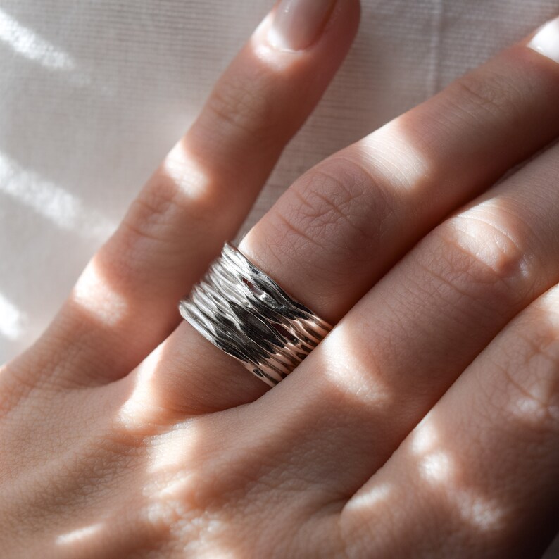 May include: A silver ring with a textured, hammered finish. The ring is stacked with several other thin bands, creating a unique and modern look.