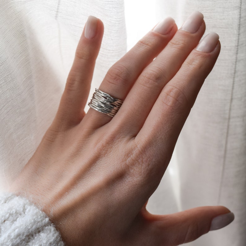 May include: A silver ring with a textured, woven design. The ring is worn on a person's finger.