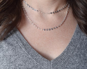 May include: A silver layered necklace with small round discs. The necklace is worn by a person with long brown hair.