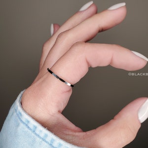 May include: A thin, black ring with silver accents is worn on a finger. The ring has a delicate design with alternating black and silver segments. The image is set against a neutral background. The brand name "BLACKBIRDLANE" is visible in the image.
