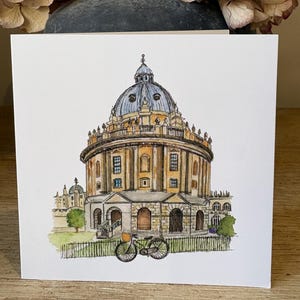 May include: A watercolour illustration of the Radcliffe Camera in Oxford, England, with a bicycle in the foreground. The card features a detailed depiction of the building's architecture and is set against a wooden surface.