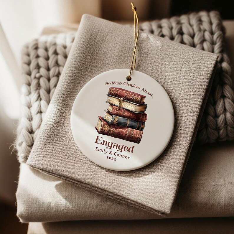 Może przedstawiać: Biała ceramiczna ozdoba ze złotym sznurkiem, z ilustracją stosu książek. Ozdoba głosi "So Many Chapters Ahead" i "Engaged" z imionami i rokiem 2025. Ozdoba spoczywa na beżowej tkaninie.