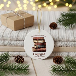 Może przedstawiać: Okrągła biała ceramiczna ozdoba ze złotym sznurkiem. Przedstawia stos książek oraz napisy "So Many Chapters Ahead..." i "Engaged Emily & Connor 2025". W tle pudełko na prezent i szyszki.