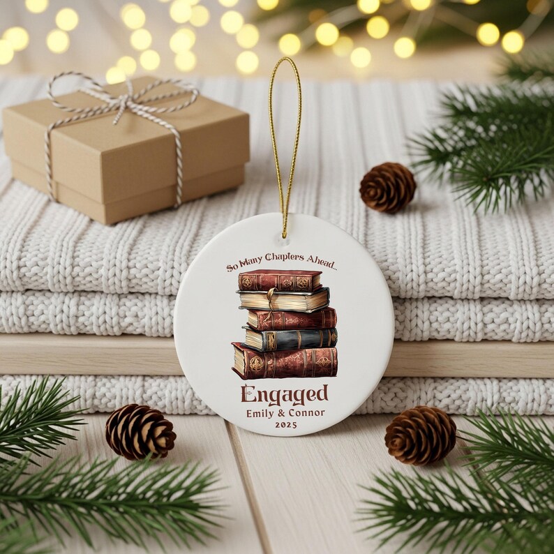 Może przedstawiać: Okrągła biała ceramiczna ozdoba ze złotym sznurkiem. Przedstawia stos książek oraz napisy "So Many Chapters Ahead..." i "Engaged Emily & Connor 2025". W tle pudełko na prezent i szyszki.