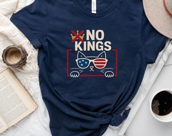 May include: A navy blue t-shirt with a graphic of a cat wearing sunglasses and a red, white, and blue American flag design. The text "NO KINGS" is printed above the cat.