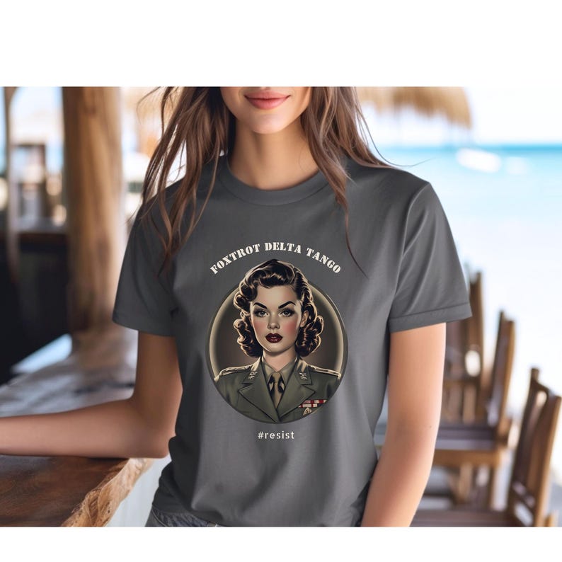 Puede incluir: Una camiseta gris antracita con un gr&aacute;fico circular blanco que presenta a una mujer con uniforme militar. El texto "FOXTROT DELTA TANGO" est&aacute; por encima del gr&aacute;fico y "#resist" por debajo.
