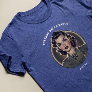 Puede incluir: Una camiseta azul con un gr&aacute;fico blanco de una mujer con uniforme militar. El gr&aacute;fico incluye el texto "FOXTROT DELTA TANGO" y "#resist".