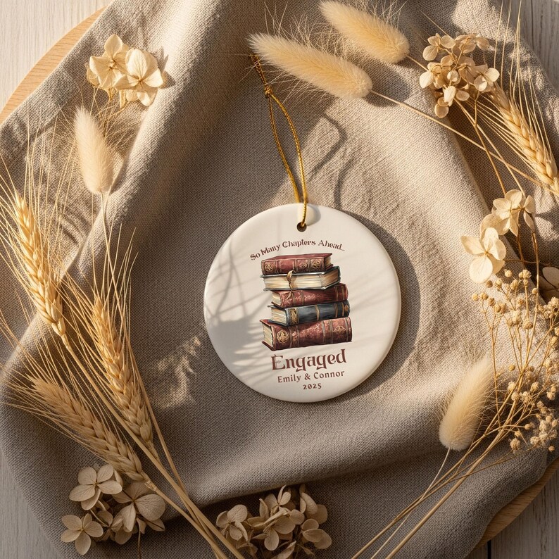 Może przedstawiać: Okrągła biała ceramiczna ozdoba ze złotym sznurkiem, przedstawiająca stos książek. Tekst brzmi "So Many Chapters Ahead" i "Engaged Emily & Connor 2025". Ozdobę otaczają suszone kwiaty i kłosy pszenicy.