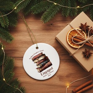 Może przedstawiać: Biała ceramiczna ozdoba z ilustracją stosu książek i napisem "So Many Chapters Ahead... Engaged Emily & Connor 2025". Ozdoba wisi na zielonym sznurku, z pudełkiem prezentowym i zielenią w tle.