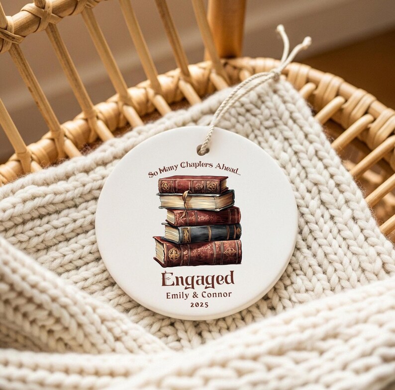 Może przedstawiać: Okrągła biała ceramiczna ozdoba z ilustracją stosu książek. Ozdoba ma napis "So Many Chapters Ahead..." na g&oacute;rze i "Engaged Emily & Connor 2025" na dole. Wisi na białym sznurku.