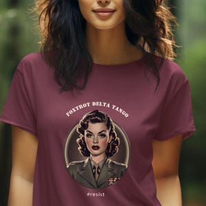 Puede incluir: Una camiseta color granate con un gr&aacute;fico blanco de una mujer con uniforme militar. El gr&aacute;fico est&aacute; rodeado por un c&iacute;rculo con el texto "FOXTROT DELTA TANGO" y el texto "#resist" debajo del gr&aacute;fico.