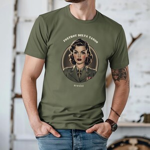 Puede incluir: Una camiseta verde con una ilustraci&oacute;n de estilo vintage de una mujer con uniforme militar. El texto "FOXTROT DELTA TANGO" est&aacute; por encima de la ilustraci&oacute;n, y el texto "#resist" est&aacute; por debajo.