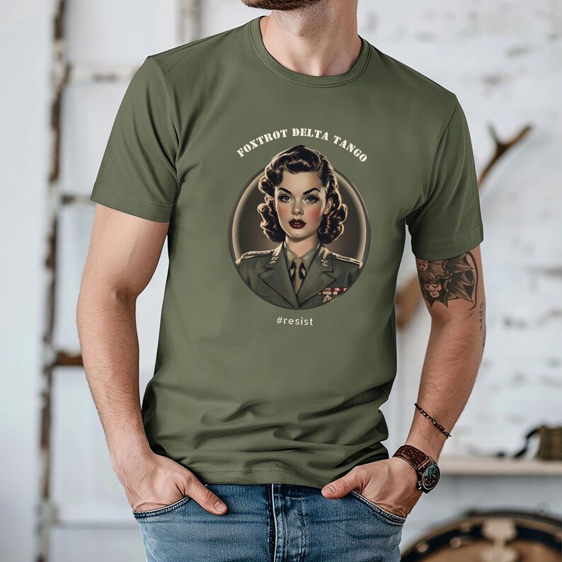 Puede incluir: Una camiseta verde con una ilustraci&oacute;n de estilo vintage de una mujer con uniforme militar. El texto "FOXTROT DELTA TANGO" est&aacute; por encima de la ilustraci&oacute;n, y el texto "#resist" est&aacute; por debajo.