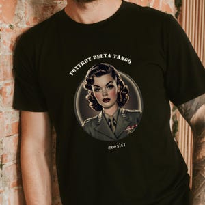 Puede incluir: Camiseta negra con un gr&aacute;fico circular blanco que presenta a una mujer con uniforme militar. El texto "FOXTROT DELTA TANGO" est&aacute; por encima del c&iacute;rculo y "#resist" est&aacute; por debajo del c&iacute;rculo.