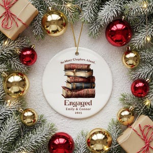 Może przedstawiać: Biała ceramiczna ozdoba świąteczna z ilustracją stosu książek. Ozdoba głosi "So Many Chapters Ahead... Engaged Emily & Connor 2025". Otoczona złotymi i czerwonymi bombkami oraz gałązkami iglastymi.