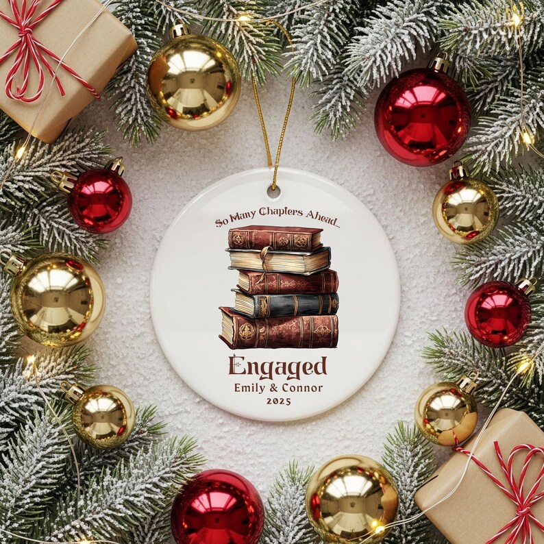 Może przedstawiać: Biała ceramiczna ozdoba świąteczna z ilustracją stosu książek. Ozdoba głosi "So Many Chapters Ahead... Engaged Emily & Connor 2025". Otoczona złotymi i czerwonymi bombkami oraz gałązkami iglastymi.