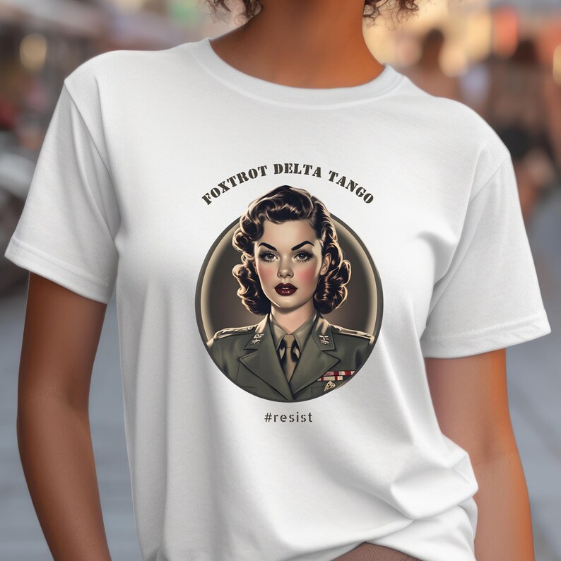 Puede incluir: Camiseta blanca con un gr&aacute;fico de una mujer con uniforme militar. El gr&aacute;fico est&aacute; en un c&iacute;rculo con el texto "FOXTROT DELTA TANGO" encima del c&iacute;rculo y "#resist" debajo del c&iacute;rculo.