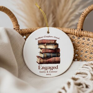 Może przedstawiać: Biała ceramiczna ozdoba w kształcie koła z ilustracją stosu książek. Ozdoba zawiera tekst "So Many Chapters Ahead..." oraz "Engaged Emily & Connor 2025". Do zawieszenia dołączono złoty sznurek.