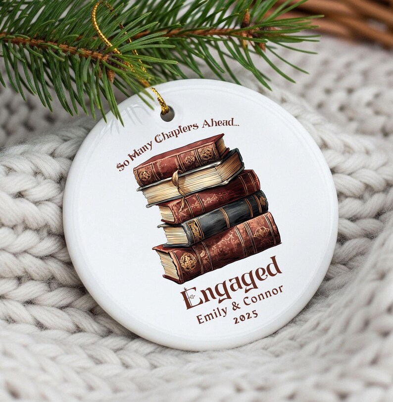 Może przedstawiać: Okrągła biała ceramiczna ozdoba ze stosu ilustrowanych książek. Tekst brzmi "So Many Chapters Ahead..." i "Engaged Emily & Connor 2025". Do zawieszenia dołączono złoty sznurek.