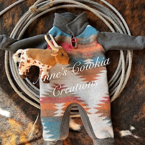 Könnte beinhalten: Ein Baby-Einteiler mit einem südwestlichen Muster in Grau, Blau, Orange und Rot. Der Anzug hat graue Ärmel und einen Kragen. Ein Stofftier-Longhorn befindet sich neben dem Outfit. Der Text "Anne's Cowkid Creations" steht auf dem Outfit.