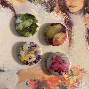 May include: Four round button badges with different floral and fruit designs. The badges feature a green leafy design, a peach and pear design, a floral design with purple and yellow flowers, and a pink flower design.