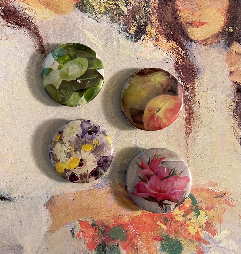 May include: Four round button badges with different floral and fruit designs. The badges feature a green leafy design, a peach and pear design, a floral design with purple and yellow flowers, and a pink flower design.