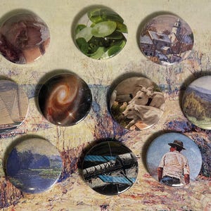 May include: Nine round, colourful buttons with various images, including a sailboat, a galaxy, a landscape, a church, a plant, a group of cows, a man in a hat, and a blue and black abstract design.