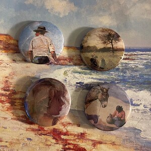 May include: Four round button badges featuring different images. The first badge depicts a man in a hat and a white shirt standing on a beach. The second badge shows a landscape with a tree in the background. The third badge has a painting of a woman's face. The fourth badge shows a woman sitting on the ground with a donkey standing nearby.