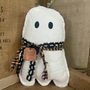 May include: A white quilted fabric ghost ornament with two black oval eyes. The ghost is adorned with a black lace ribbon and a tan and black checkered fabric tie. A small square copper-colored tag hangs from the ribbon with the text Harvest Blooms.