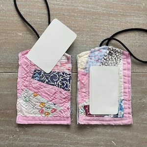 May include: Two handmade fabric badge holders with a patchwork design. The holders are primarily pink with patches of blue, white, and patterned fabric. Each holder has a white card and a black lanyard.