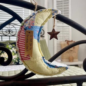 May include: A yellow, blue, and red patchwork crescent moon with a brown star hanging from it. The moon has a brown tag attached that says "I love you to the moon + back".