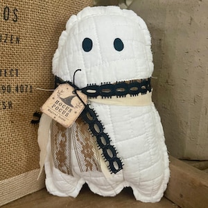 May include: A white quilted fabric ghost figure with two black circular eyes. It is adorned with a black lace trim, a beige ribbon, and a small rectangular tag featuring a witch hat illustration and the text Hocus Pocus Magical Goods.