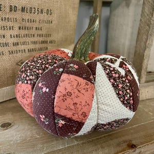 May include: A fabric pumpkin decoration featuring a patchwork design of brown, pink, and cream-coloured cotton segments. The segments are patterned with small floral prints and polka dots, and the pumpkin is topped with a green fabric stem.