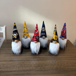 May include: Six decorative gnomes with white beards and fabric hats. The hats feature logos and colors of various hockey teams, including black and gold, red and black, and blue and gold. They are arranged on a wooden surface.