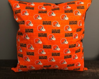 May include: Orange and brown throw pillow with a repeating pattern of the Cleveland Browns logo. The logo features a football helmet with the words "Cleveland Browns" written below it.