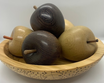 May include: A wooden bowl with a decorative design on the bottom, filled with four wooden apples. Two of the apples are dark brown and two are light brown.