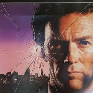 May include: A close-up of a man's face with a cracked glass pane over his eye. The glass has a bullet hole in it. The background is a cityscape at night.