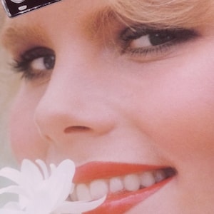 May include: A close-up of a woman's face with a silver number plate that says "STAR 80" on her forehead. She is smiling and has a white flower in her mouth.