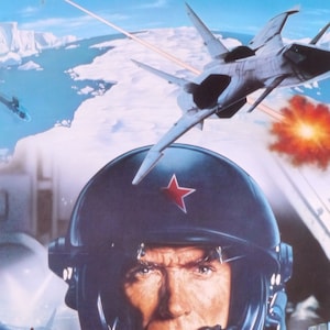 May include: A close-up of a pilot wearing a blue helmet with a red star on the front. The pilot is looking directly at the viewer. In the background, a white fighter jet flies over a snowy landscape.