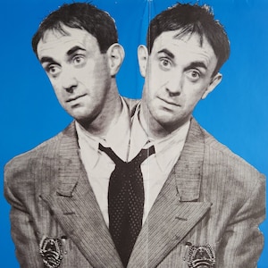 Puede incluir: Una fotografía en blanco y negro de un hombre con dos cabezas que lleva un traje gris con una corbata negra. El hombre mira a la cámara con una expresión seria. La foto está sobre un fondo azul brillante.