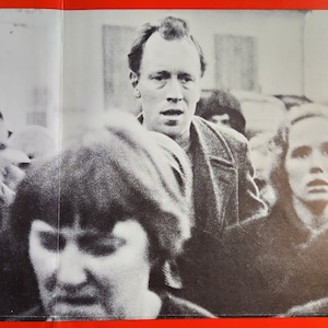 以下が含まれることがあります： カメラに向かって真剣な表情をした男性を含む、人々の白黒写真。男性は暗いジャケットを着ており、短く暗い髪をしています。写真の中の他の人々はぼやけていてピントが合っていません。