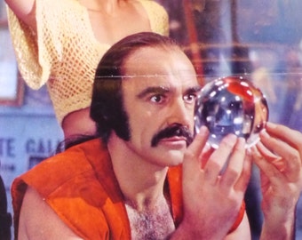 Può includere: Un uomo con i baffi e capelli lunghi e scuri guarda intensamente una sfera di cristallo. Indossa una camicia rosso-arancione.