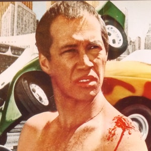 May include: A man with a bloody injury on his shoulder stands in front of a green car that has flipped over. The car is in the foreground of the image, and the man is in the background. The man is looking at the camera with a serious expression on his face.