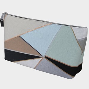 May include: A geometric pattern cosmetic bag with a silver zipper. The bag features a design of triangles in shades of gray, beige, black, and blue-green, with thin gold-colored lines separating the shapes. The bag is rectangular and appears to be made of fabric.