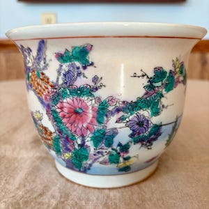 May include: A white ceramic planter with a flared rim, adorned with a floral design in pink, purple, green, and orange hues. The planter features a light blue base and a delicate, hand-painted aesthetic.