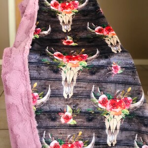 May include: A soft pink faux fur throw blanket with a floral skull pattern on the front. The pattern features a white skull with horns and red, pink, and white flowers. The background is a dark brown woodgrain pattern.