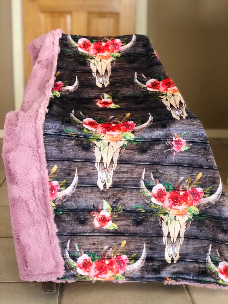 May include: A soft pink faux fur throw blanket with a floral skull pattern on the front. The pattern features a white skull with horns and red, pink, and white flowers. The background is a dark brown woodgrain pattern.